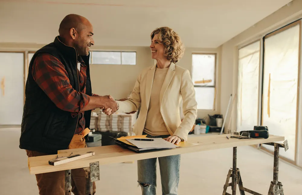 Professional general contractor in work attire with tool belt consulting with homeowner in front of suburban home, representing trusted home improvement and remodeling services for local TV advertising.